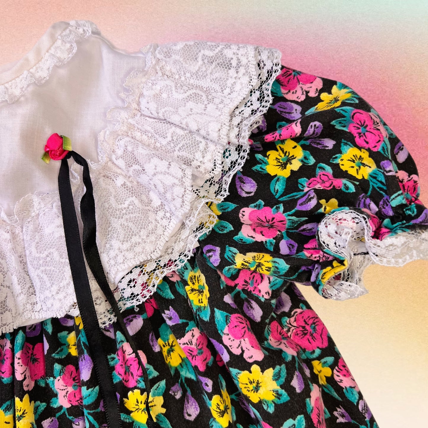 a very 80's white ruffle collar with hot pink rosette attached to a falling black ribbon. A cap sleeve with lace trim, with a black cotton material background amid a vibrant magnolia pattern of pinks, purples, yellows, and green. 